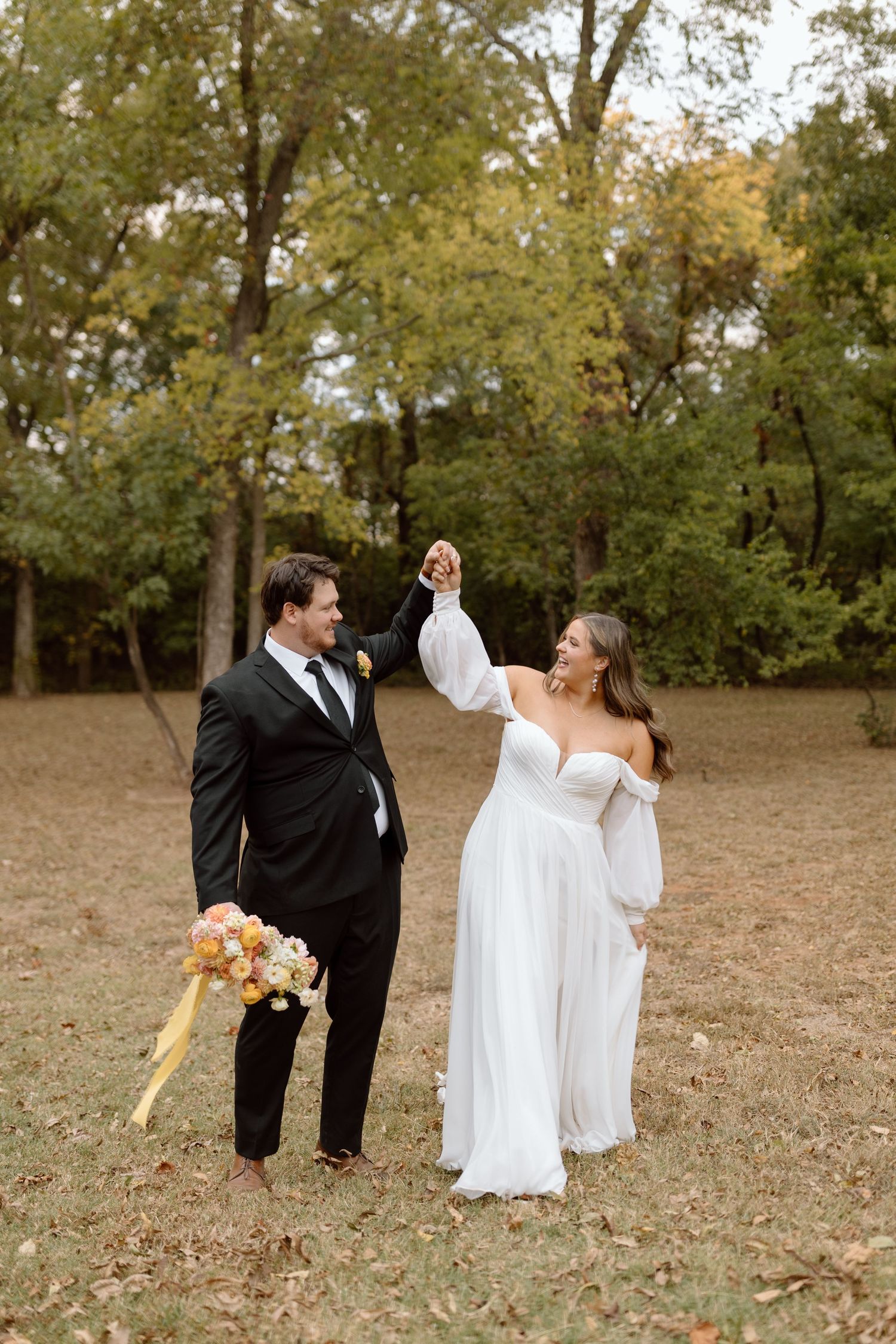 Couple in elegant wedding attire dancing and twirling together in a scenic outdoor autumn setting.