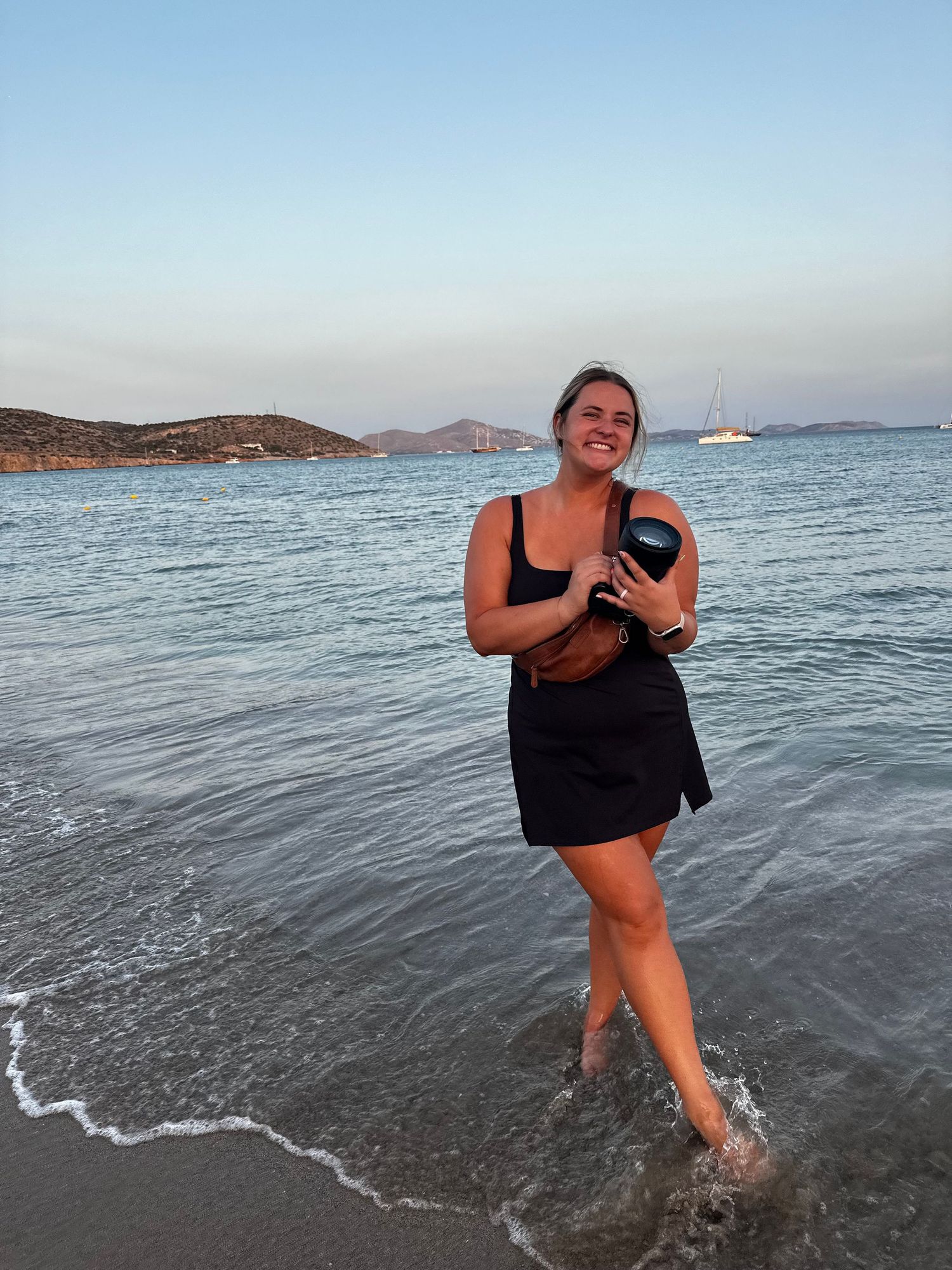 A beachgoer stands joyfully in shallow ocean waves at sunset with mountains visible in the background.