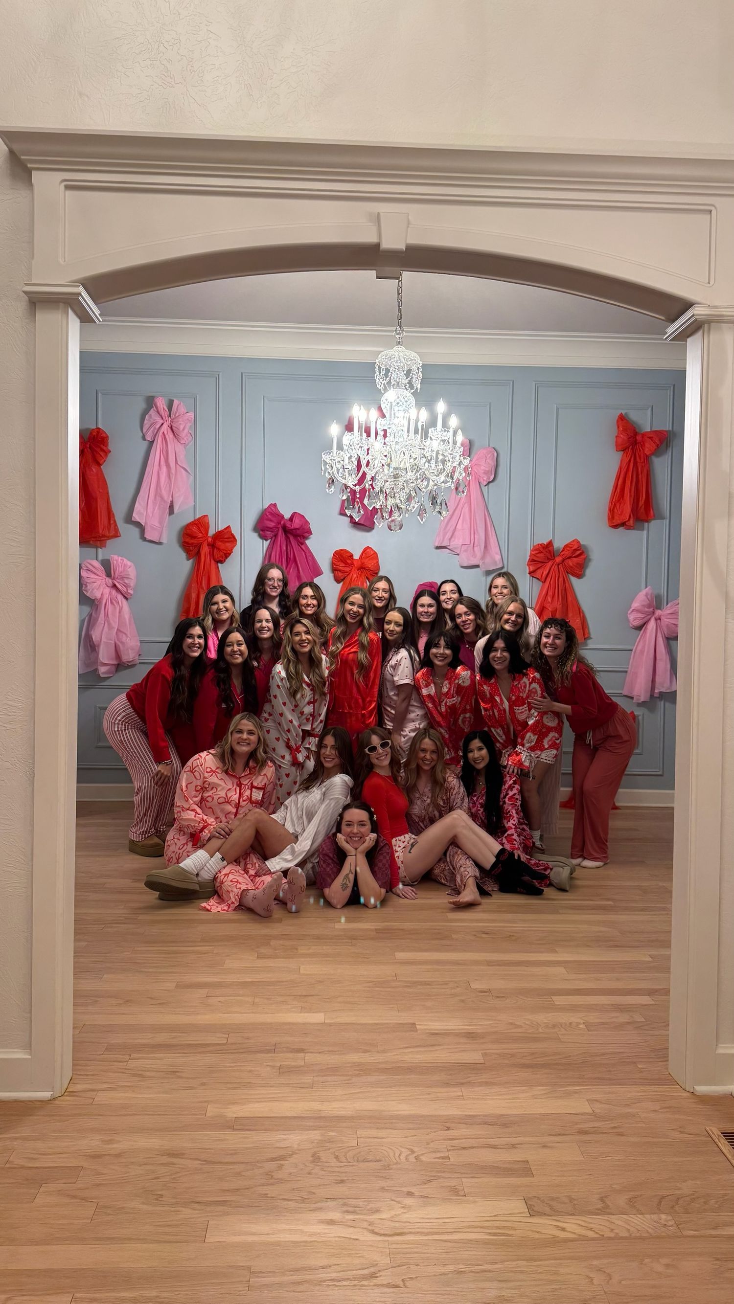 A group poses in a room decorated with red and pink bows on the walls beneath an elegant crystal chandelier.
