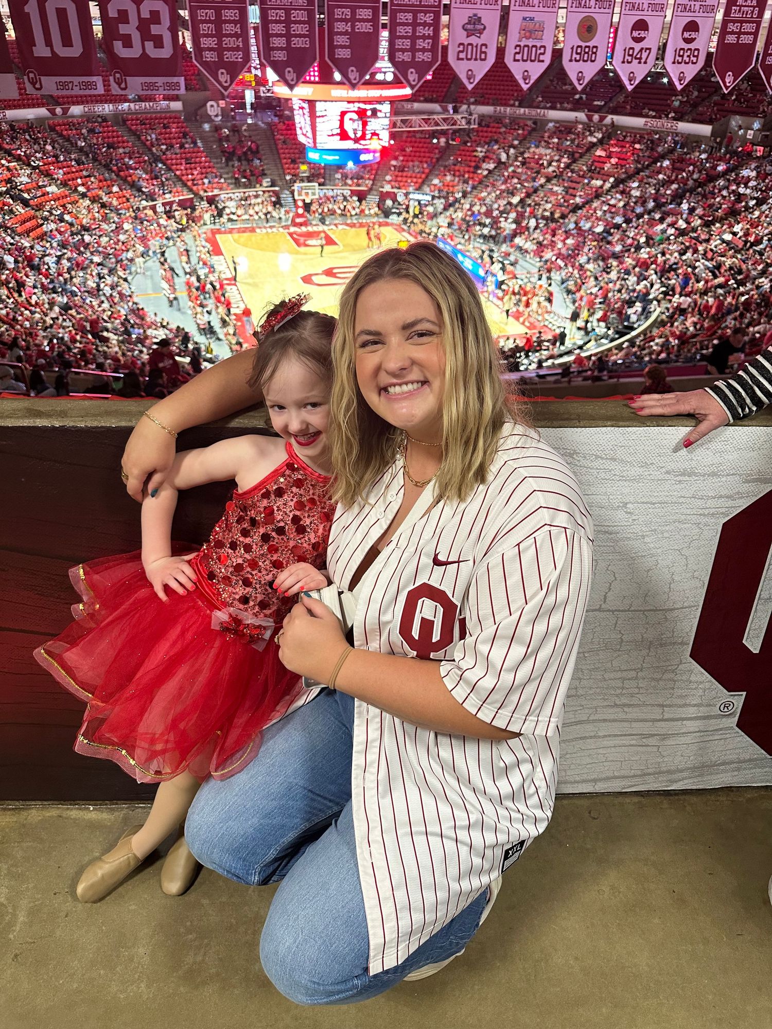 A person wearing an Oklahoma Sooners baseball jersey poses alongside a red tutu at a stadium event.