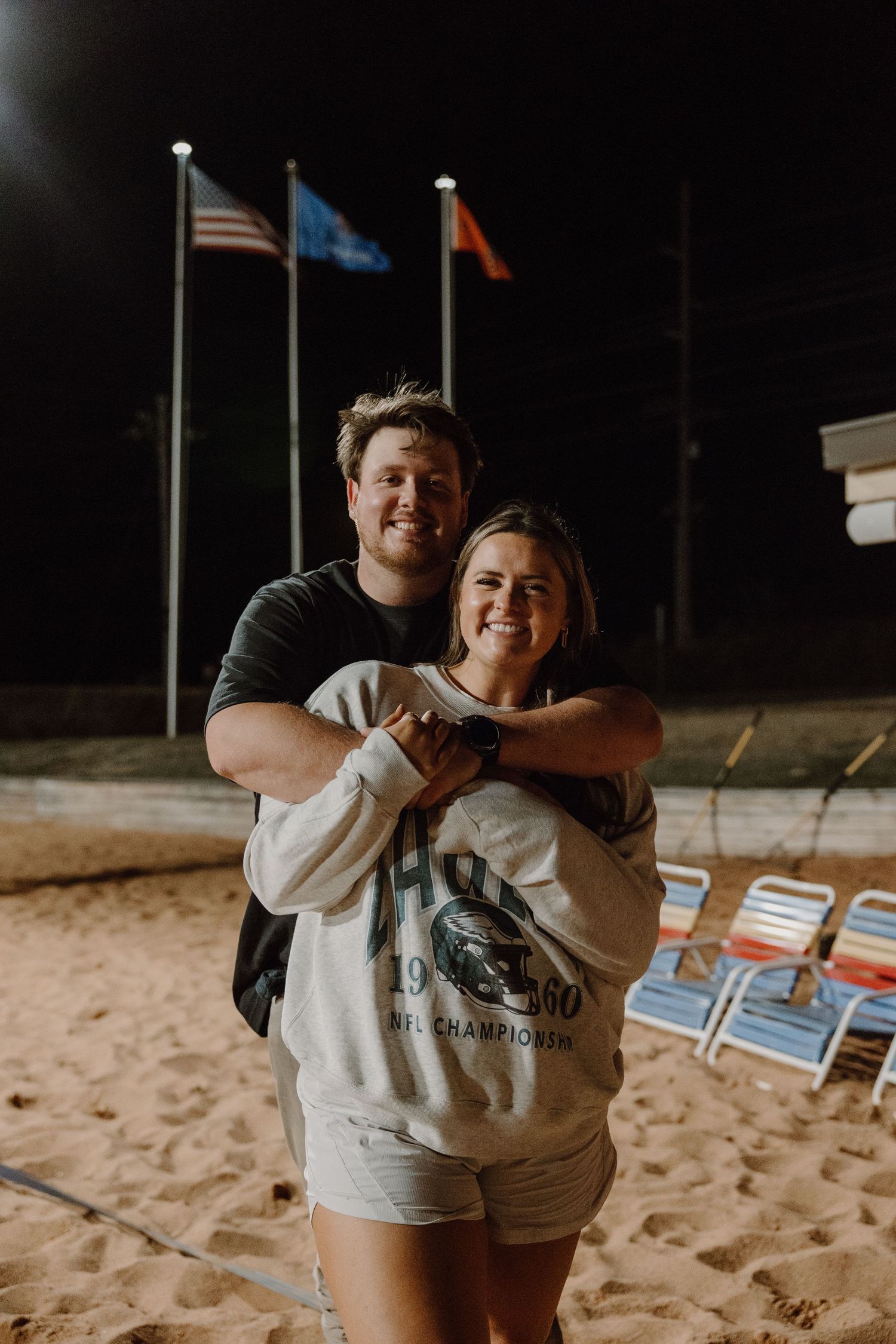 Couple embracing on beach at night with flags in background.