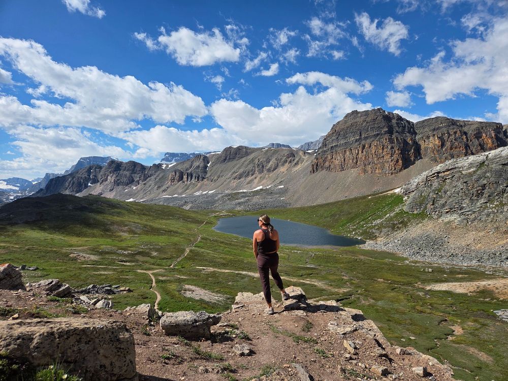 Hikers traverse a scenic mountain trail with dramatic rocky peaks and green valleys under bright blue skies.