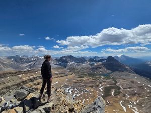 A silhouetted hiker stands on a mountain peak overlooking a dramatic alpine landscape with winding roads below.