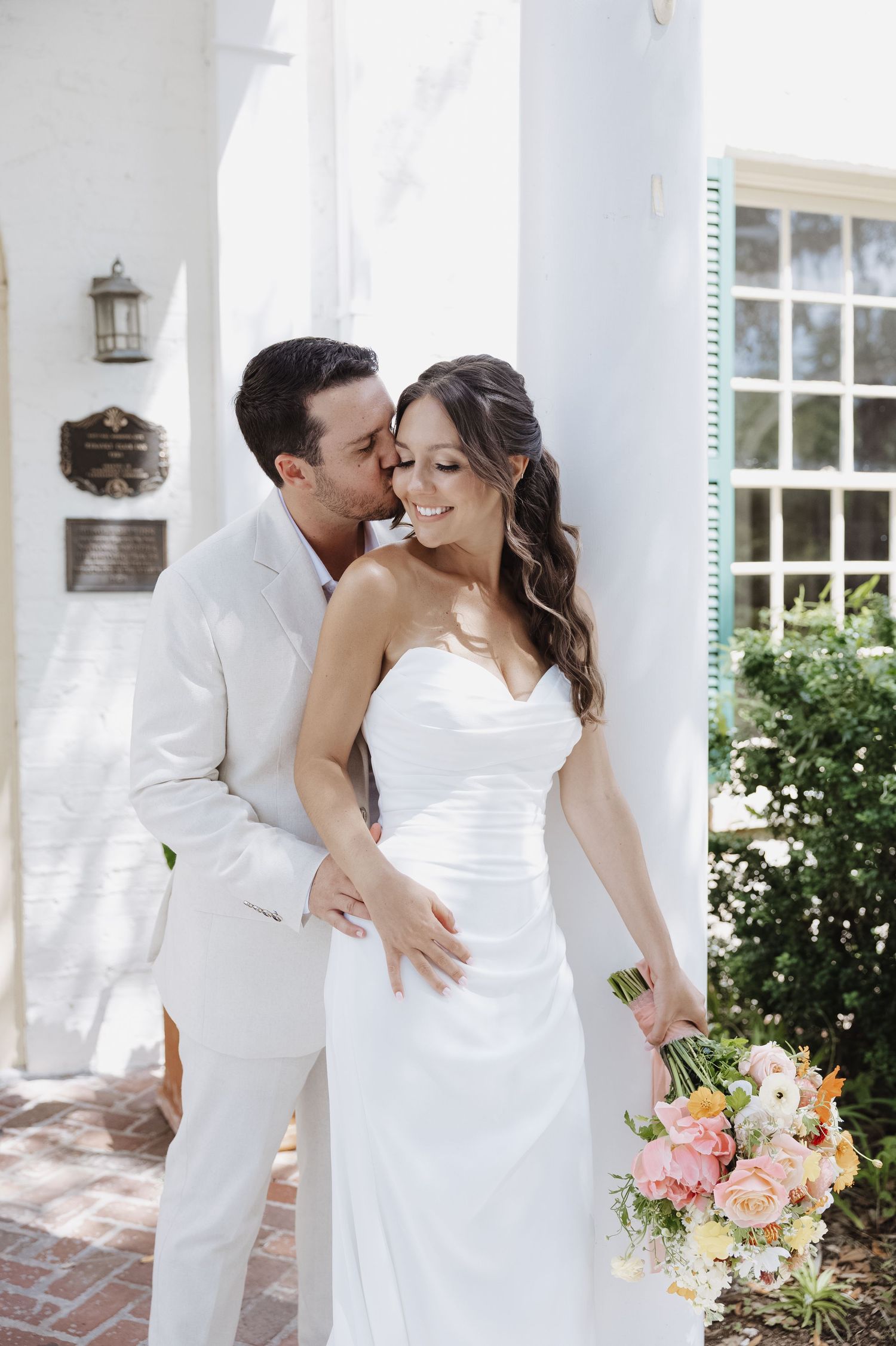 Groom in white suit kissing bride on cheek as she smiles holding colorful wedding bouquet.