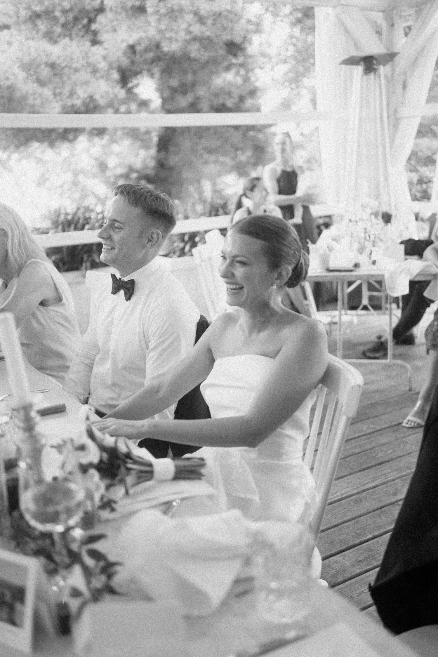 A joyous black and white candid moment of laughter between wedding guests at a reception in a rustic barn setting.