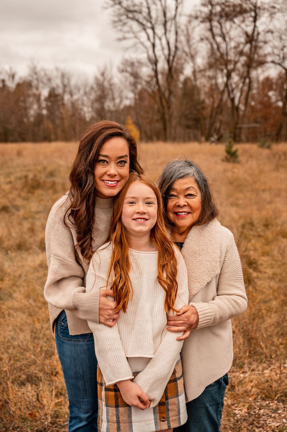 Three generations of family members share joyful moments in a rustic autumn field wearing cream-colored sweaters.