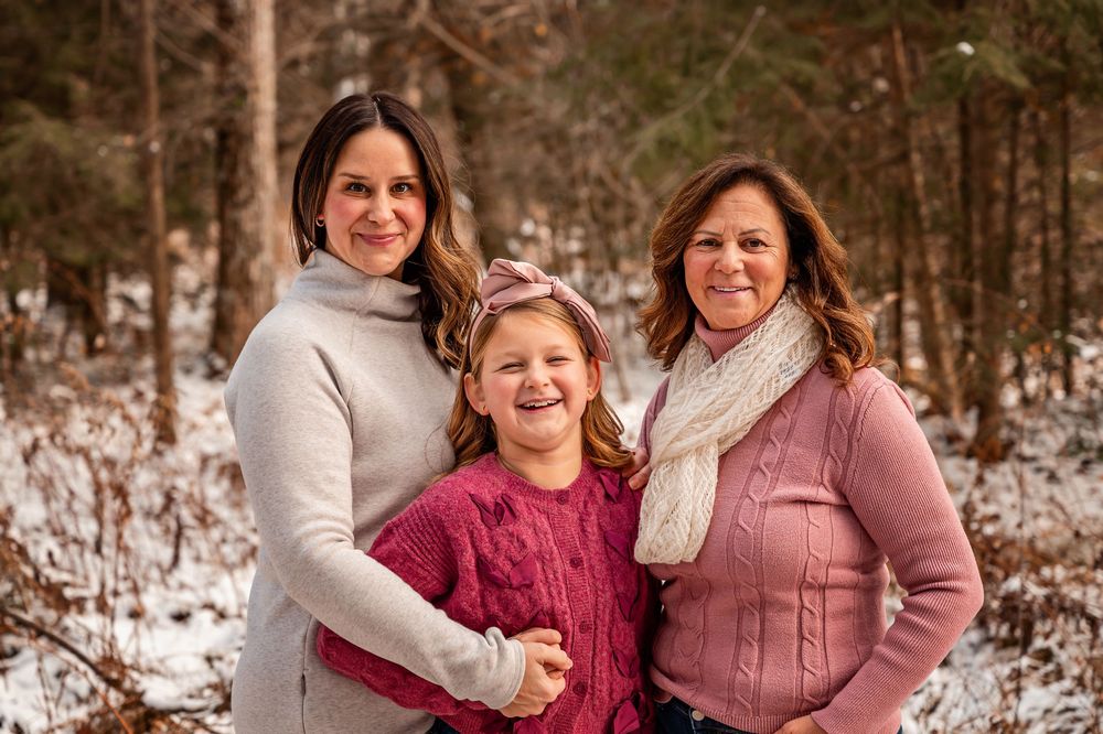 Three people wearing winter sweaters and scarves pose together in a snowy forest setting.