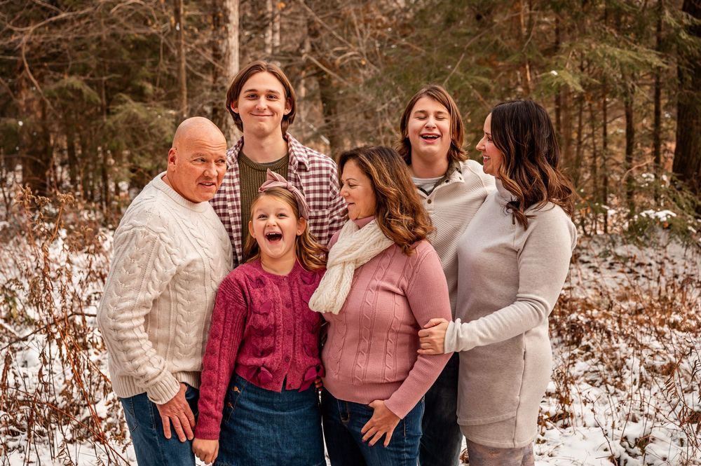 A family of six poses together outdoors in winter, wearing cozy sweaters and sharing laughter in a snowy forest setting.