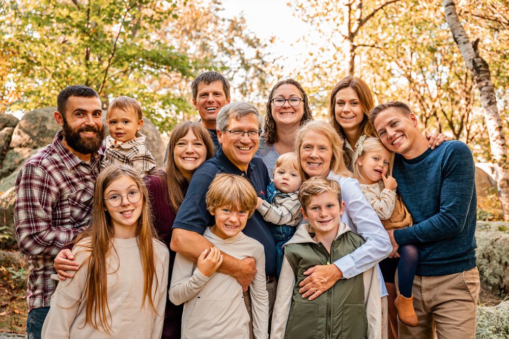 Extended family gathering poses for a group photo in an autumn setting with fall colors.