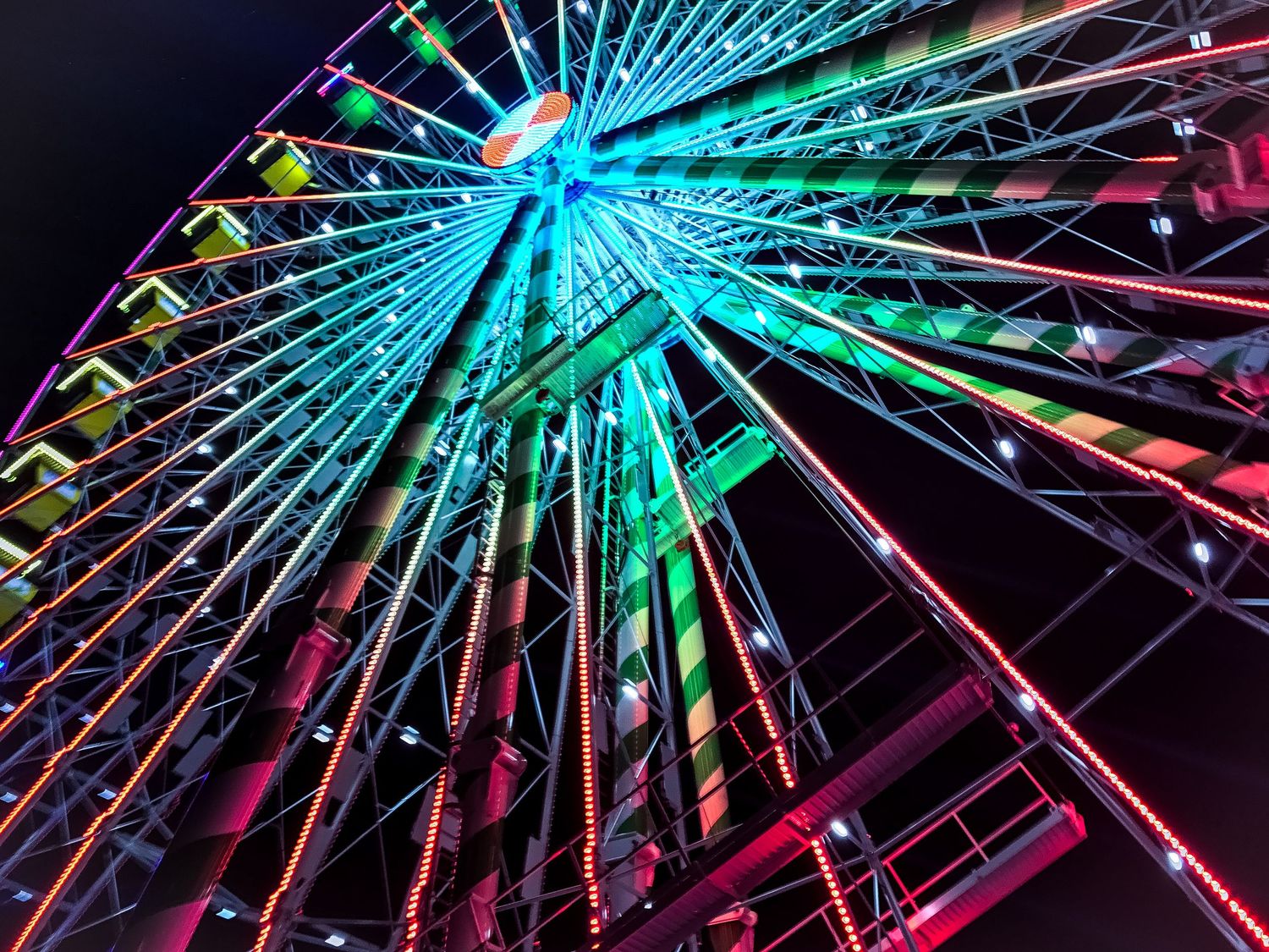 Illuminated ferris wheel glowing with vibrant blue, green and pink lights against a dark night sky.