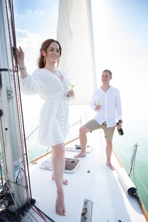 Couple celebrating on a sailboat, woman in white dress holding champagne glass with man pouring bottle in background.