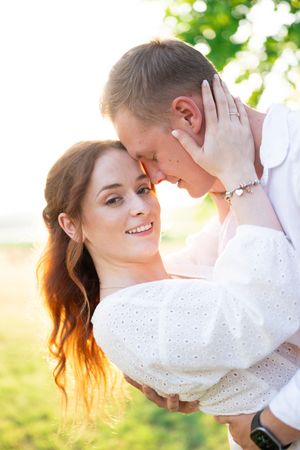 Smiling woman in white dress gazes up at man as they embrace in warm sunlight.