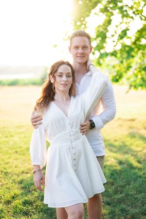 Couple in white outfits embracing in golden sunlight near green tree in open field.