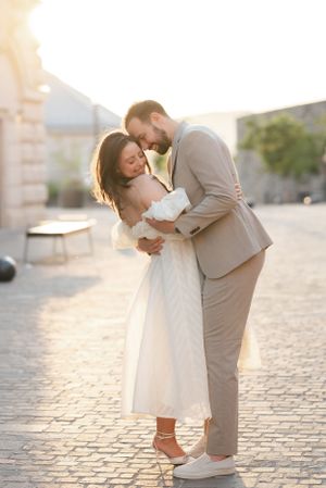 A couple embracing romantically in golden sunset light on a cobblestone square, bride in white midi dress.