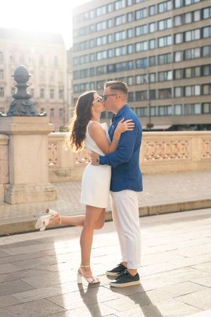 Couple sharing a romantic kiss in a sunny city square, woman in white dress, man in blue jacket.