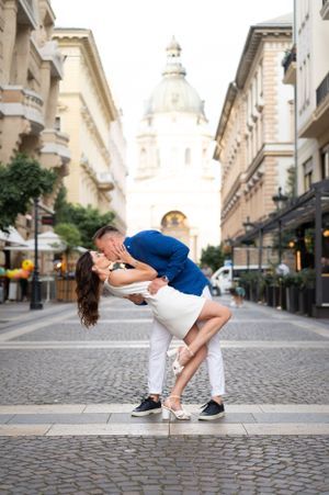 Couple sharing a romantic dip kiss on a cobblestone street with a domed cathedral in Budapest.