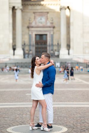Couple embracing and kissing in a sun-drenched European plaza with a grand classical building behind.