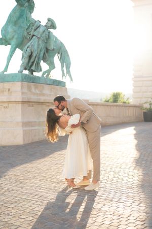 Couple sharing passionate kiss as groom dips bride near bronze equestrian statue in golden sunlight.