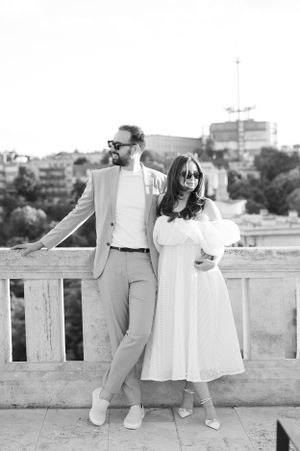 Stylish couple posing in black and white photo on a terrace overlooking a European cityscape.