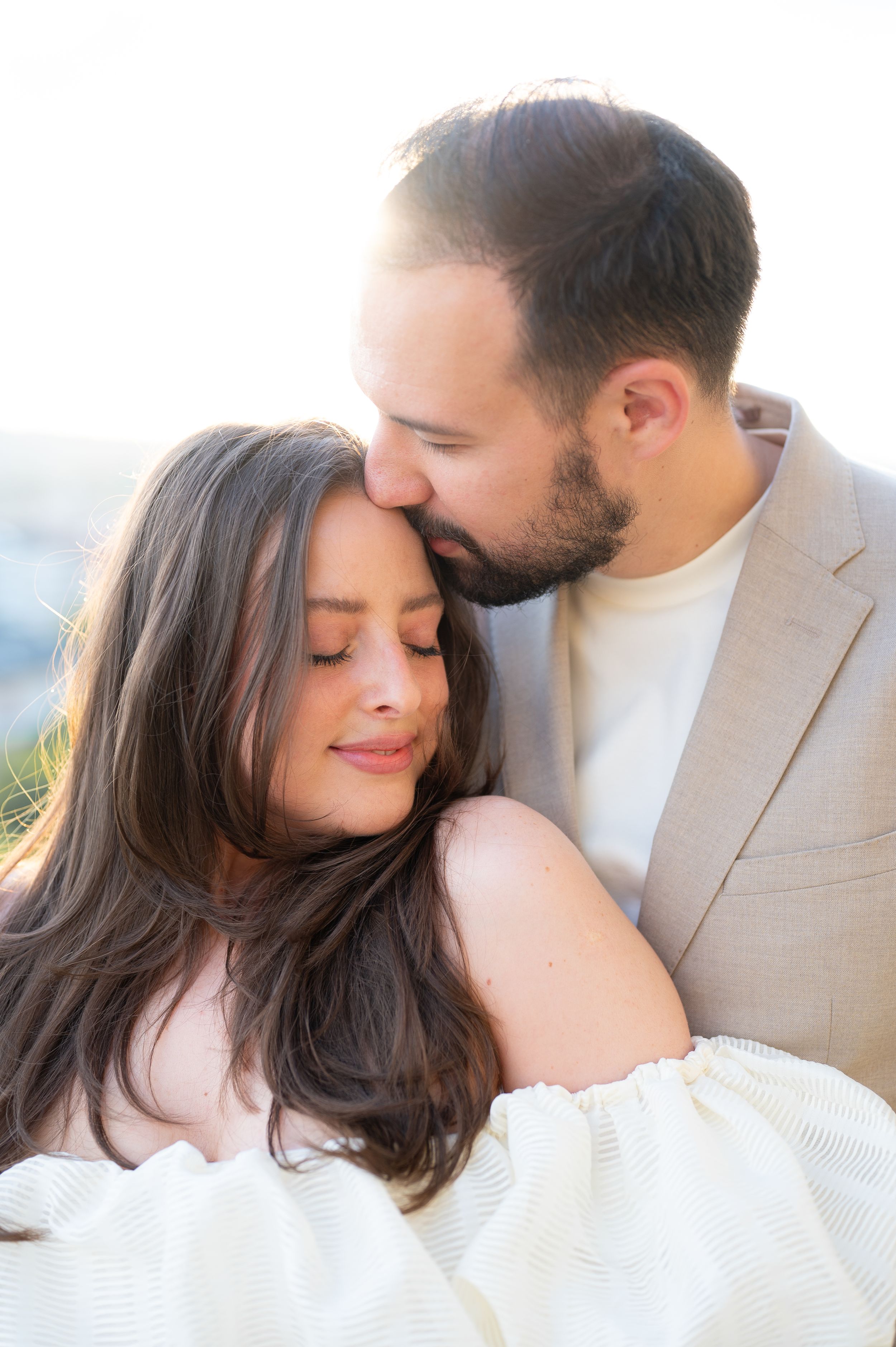 Couple sharing a tender moment, man kissing woman's forehead in golden sunlight during engagement shoot.