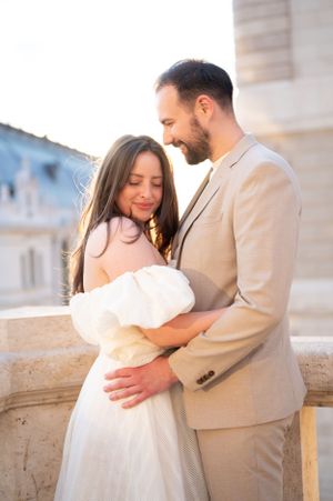 Groom in beige suit embracing smiling bride in white dress on sunlit European terrace.