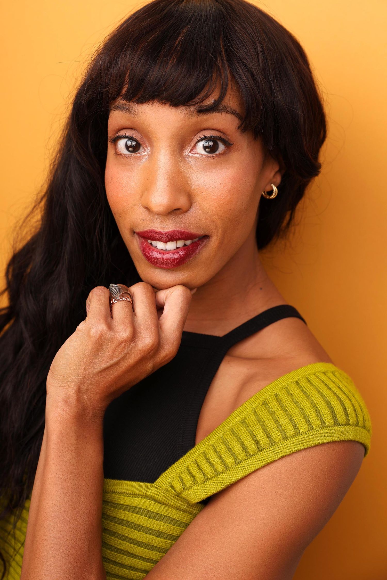 Professional beauty portrait with long black wavy hair wearing an off-shoulder olive top against brown background.