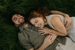Happy couple lying together on green grass while laughing and embracing in casual summer attire.
