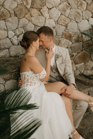 A romantic wedding couple embraces against a rustic stone wall with palm frond shadows creating a tropical ambiance.