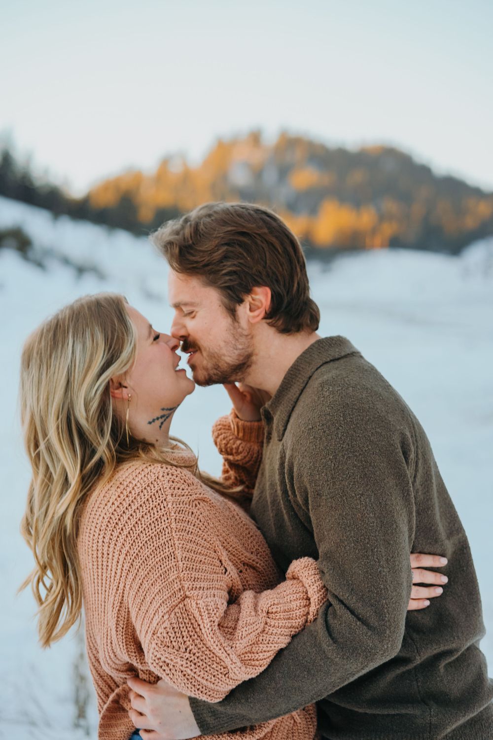 A couple in cozy sweaters share an intimate moment against a snowy mountain backdrop with autumn trees.