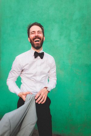 Professional portrait against green backdrop shows person in white dress shirt and bowtie with gray pants.
