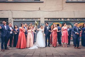 Wedding party in formal attire poses in front of rustic building facade with bridesmaids in coral dresses.