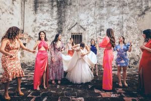 A panoramic wedding party photo showing a group in coral and floral dresses posing against a rustic stone wall.
