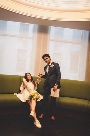 A couple in formal attire sits on a curved green couch in an elegant room with soft lighting.