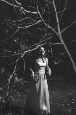 A ghostly figure in a white dress stands among dark bare tree branches in an eerie black and white photograph.