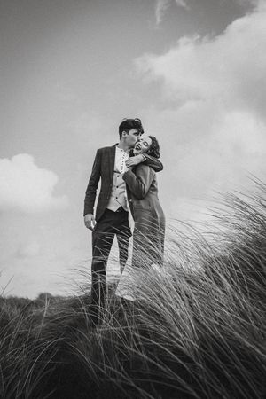 A romantic black and white photograph of a couple embracing on a grassy hill with tall grass swaying in the wind.