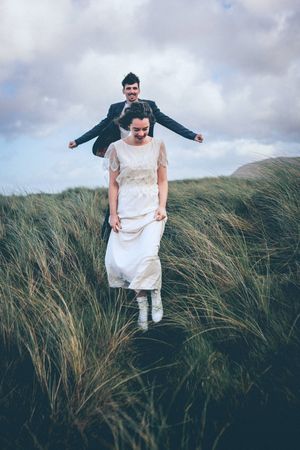 Two people stand among tall grass on a hillside with cloudy skies, one wearing a white dress and another in dark clothing.