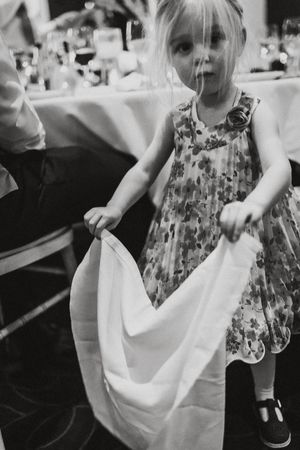 A young child in a floral dress plays with a white napkin at a dining table.