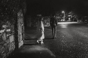 A black and white nighttime photograph showing two figures walking a dog on a dimly lit street.