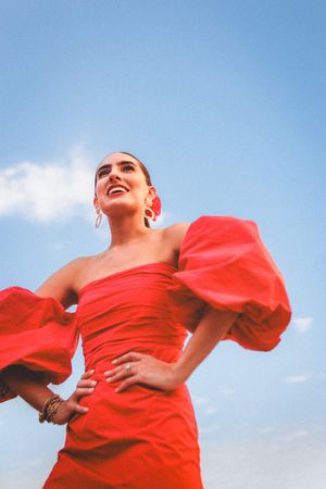 A person in a flowing red dress twirls joyfully against a bright blue sky with white clouds.