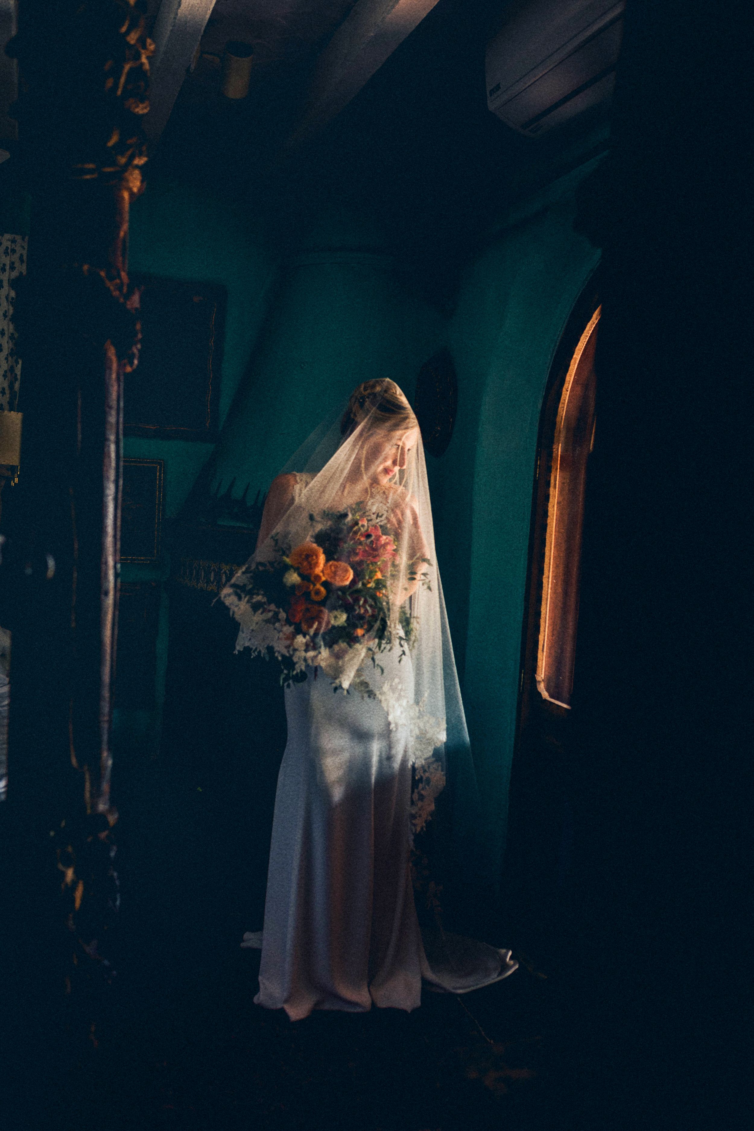 A bride in a flowing white dress and veil stands in a dark church interior holding a vibrant autumn bouquet.