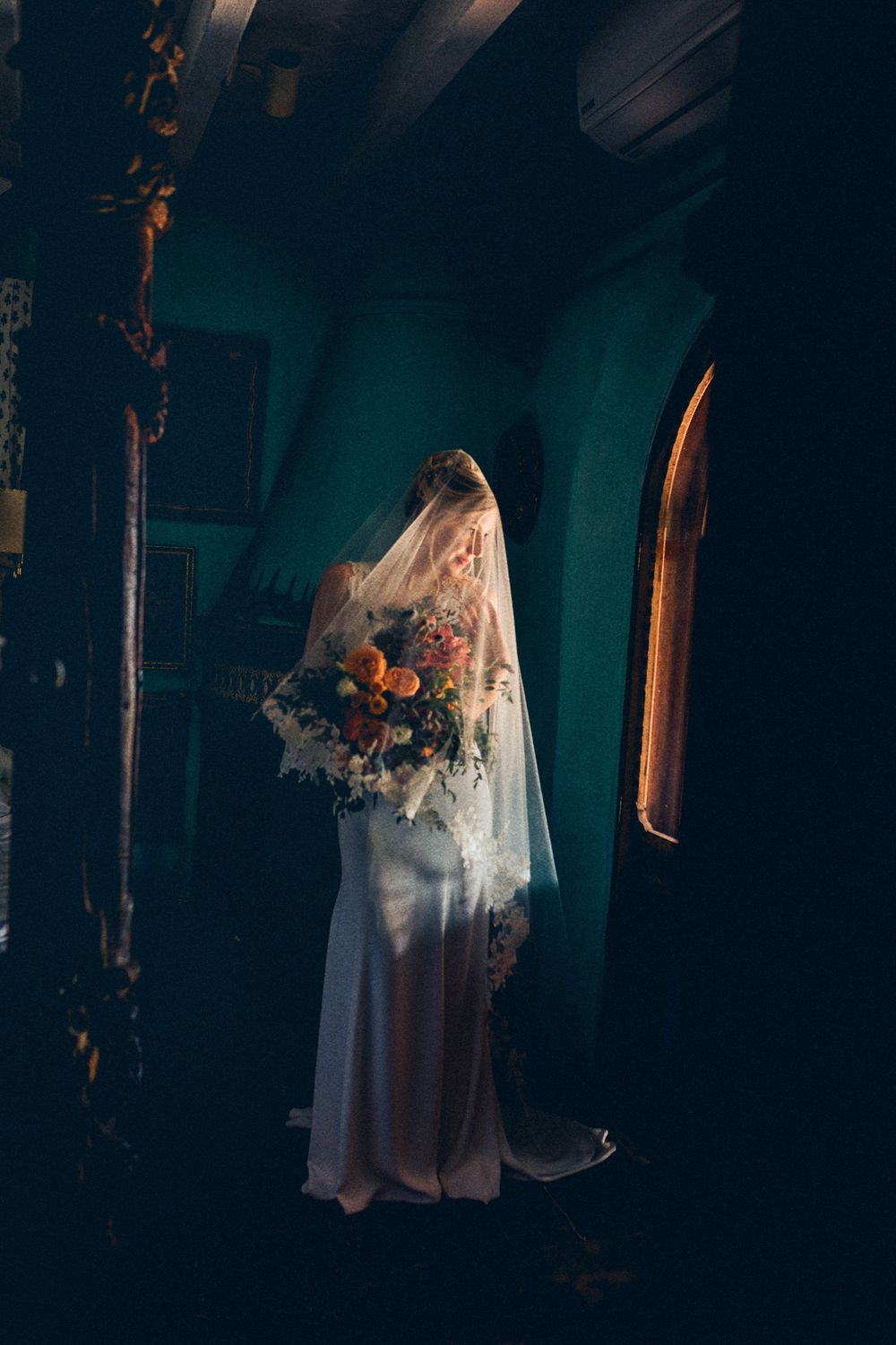 A bride in a flowing white dress and veil stands in a dark church interior holding a vibrant autumn bouquet.