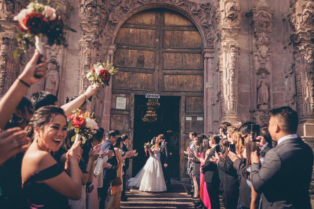 Wedding guests toss flower petals as newlyweds exit an ornate church with carved stone archway and wooden doors.