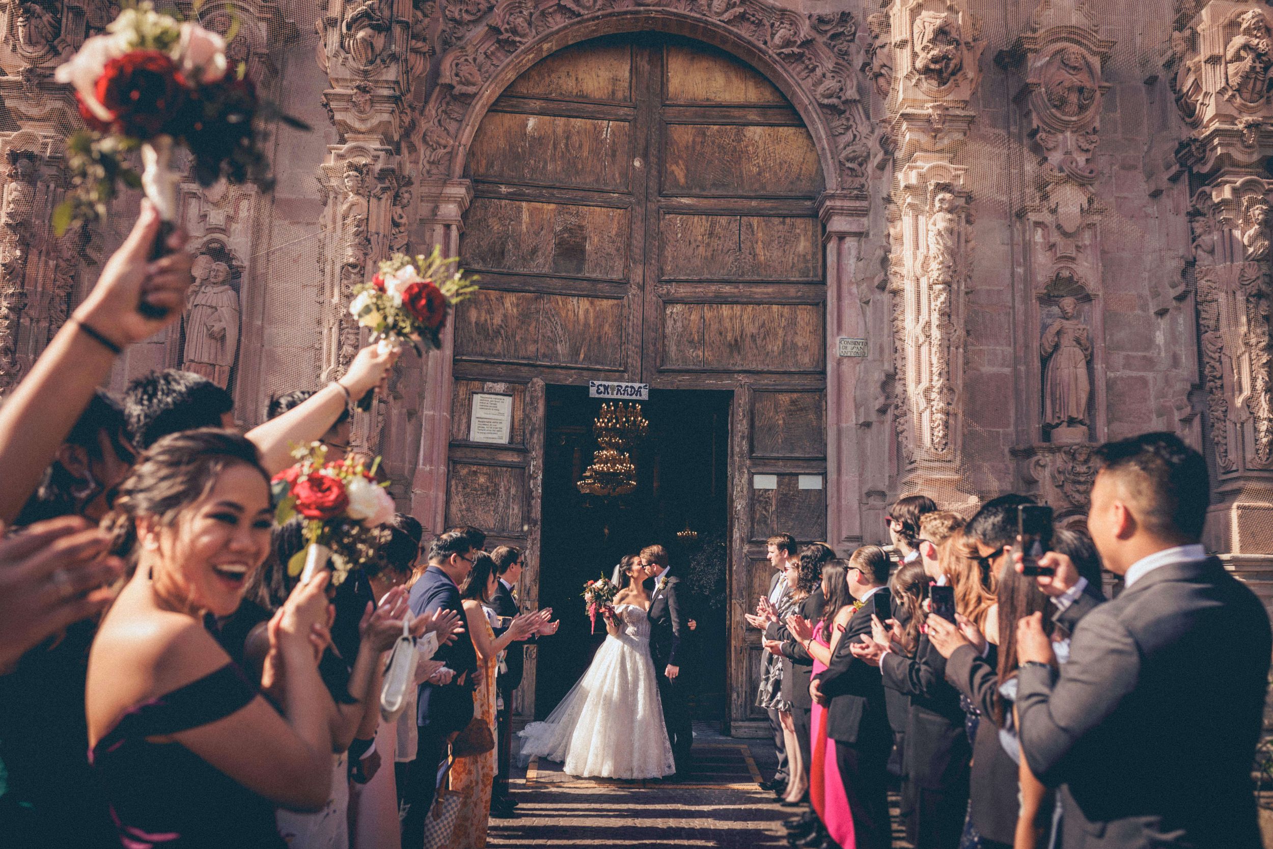 Wedding guests toss flower petals as newlyweds exit an ornate church with carved stone archway and wooden doors.