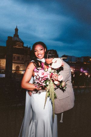 A couple shares a romantic moment during their wedding photoshoot against a dramatic evening sky with historic buildings.