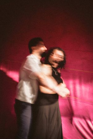 Motion blur captures two dancers performing a dramatic spin against a vibrant pink backdrop in a dance studio.