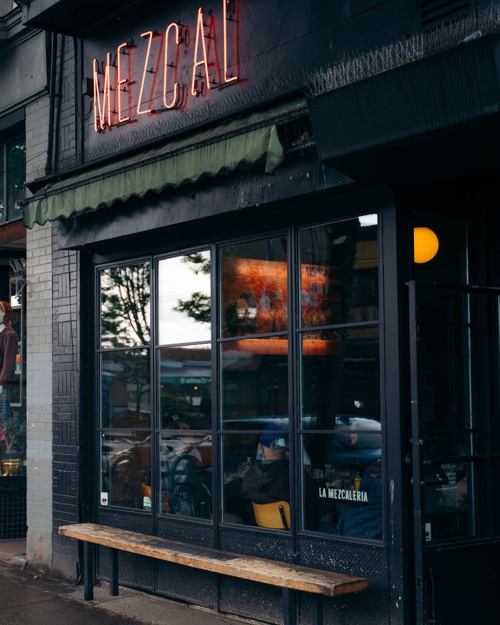 Mezcal bar exterior with neon sign, dark facade, large windows, and a wooden bench on the sidewalk.

Vancouver Canada Scene