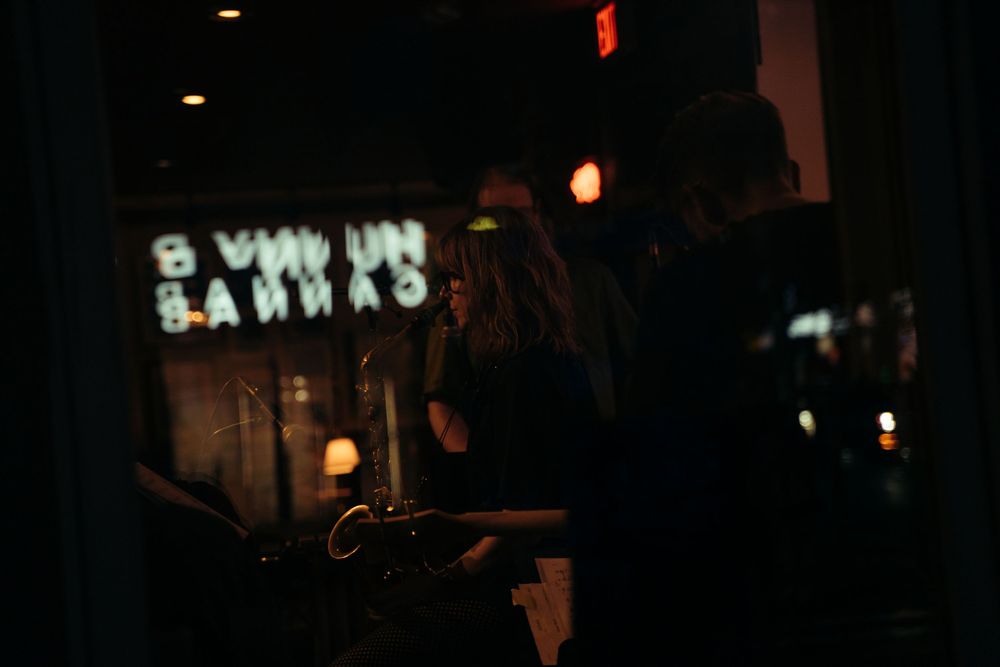 Dimly lit bar interior with patrons silhouetted against glowing neon signs reflected in the window. Vancouver Scene