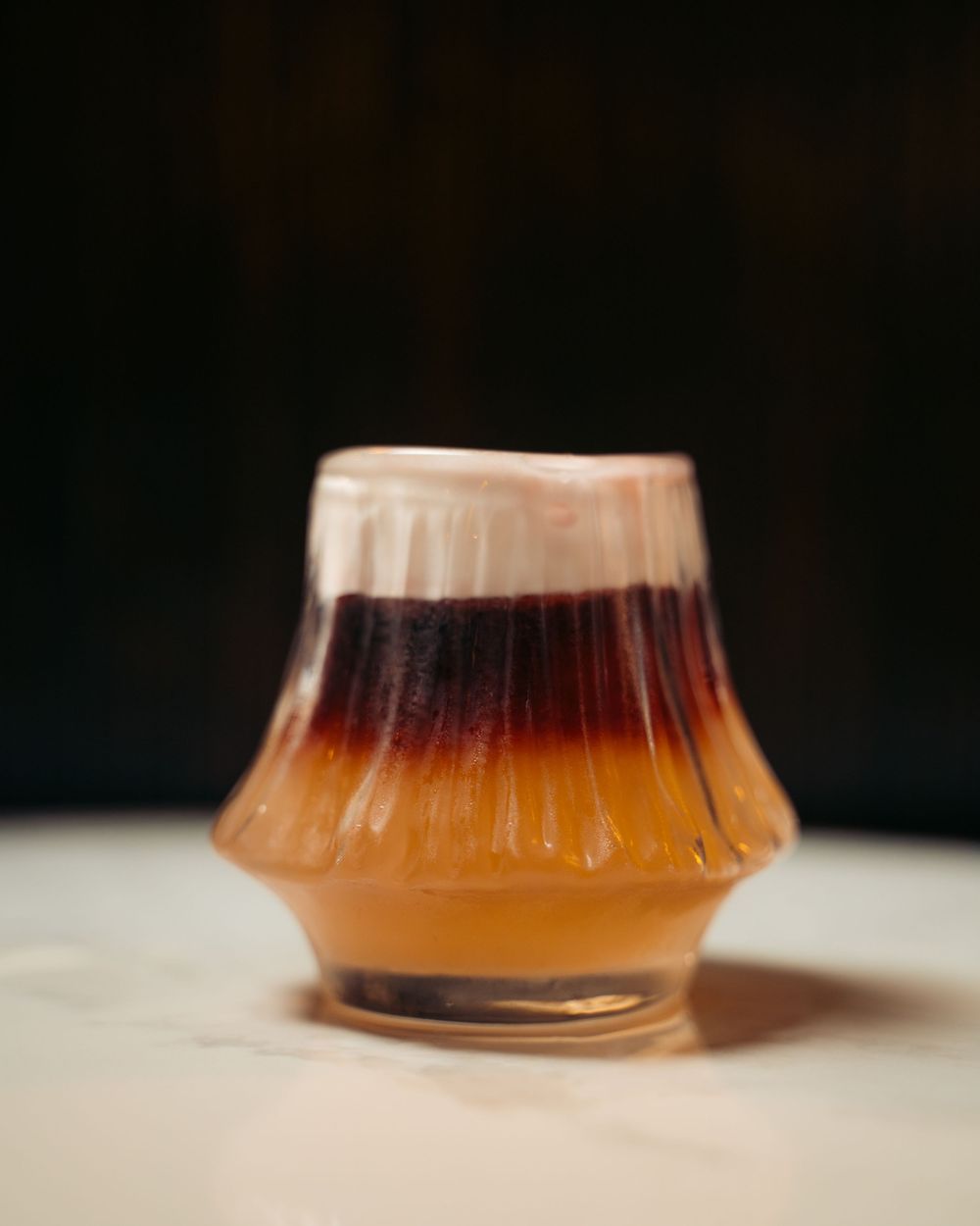 Layered brown and cream cocktail served in a unique ribbed glass on a white marble surface. Vancouver Scene