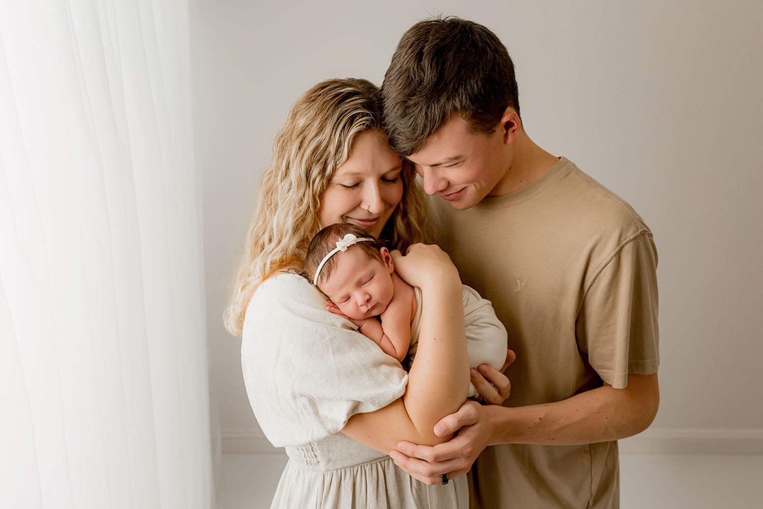 New parents smiling and holding sleeping newborn baby girl by window during family portrait session at Summerville South Carolina photograph