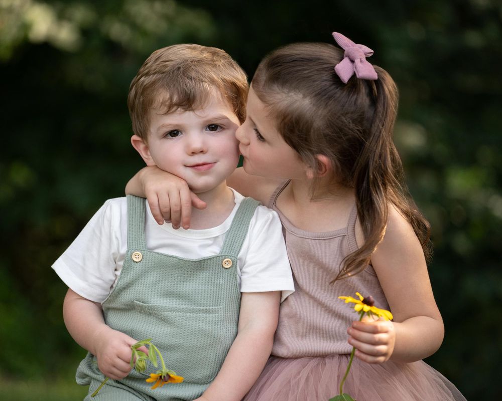 Little one wearing sage green overalls touches daisies in a gentle outdoor nature scene with green background.